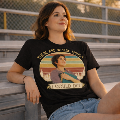Woman wearing a black t-shirt with a graphic design and text, sitting on bleachers.