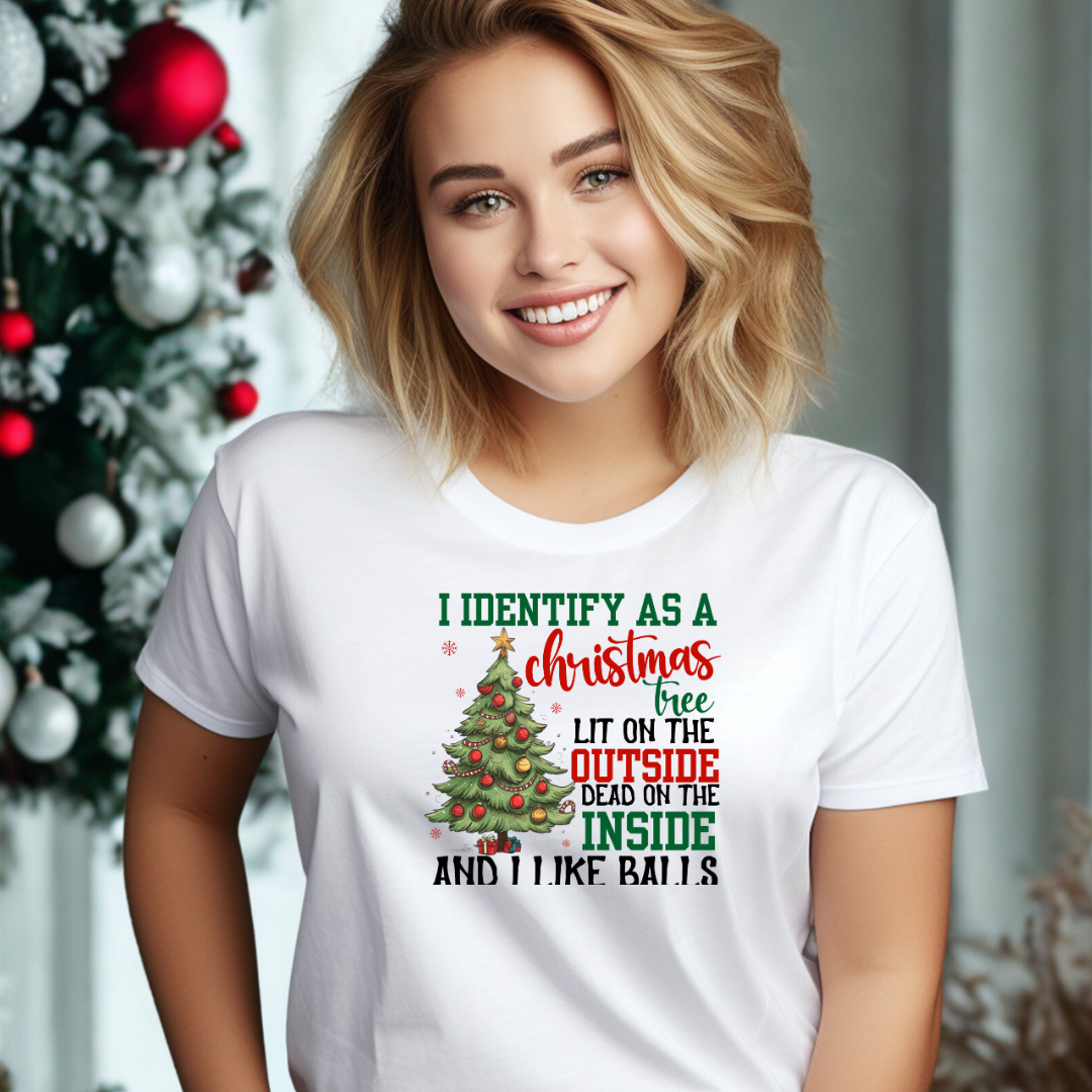 A smiling blonde woman wearing a white graphic t-shirt with a Christmas tree design and text, posed in front of a decorated Christmas tree.