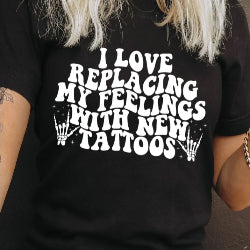 Person wearing a black t-shirt with I love replacing my feelings with new tattoos white text design.