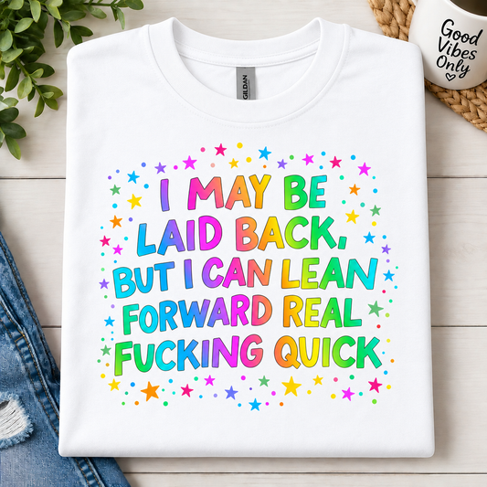 A white folded graphic T-shirt with colorful rainbow text reading 'I MAY BE LAID BACK, BUT I CAN LEAN FORWARD REAL FUCKING QUICK' surrounded by small multicolored stars, styled on a light wood surface with jeans, greenery, and a woven mat.