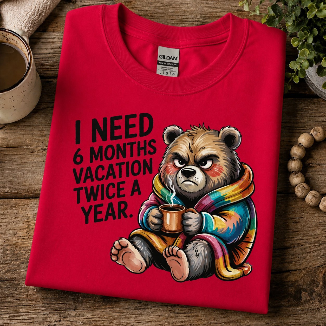 A folded heather red Gildan t-shirt with a humorous graphic of a grumpy cartoon bear wrapped in a blanket and holding a mug, next to the text 'I NEED 6 MONTHS VACATION TWICE A YEAR.' The shirt is styled on a rustic wooden table with a mug, plant, and beads.