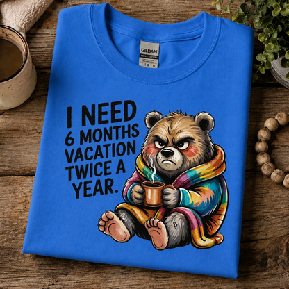 A folded heather royal Gildan t-shirt with a cartoon bear graphic and the text 'I NEED 6 MONTHS VACATION TWICE A YEAR.' displayed on a rustic wooden table with lifestyle props.