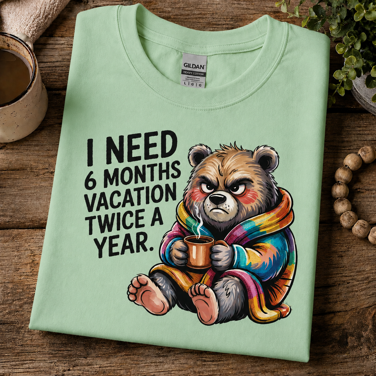 A folded mint green Gildan t-shirt with a humorous graphic of a grumpy bear in a rainbow blanket holding a mug, with the text 'I NEED 6 MONTHS VACATION TWICE A YEAR.' on a rustic wooden background.