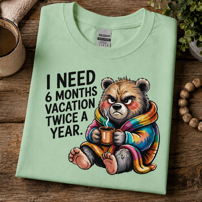 A folded mint green Gildan t-shirt with a humorous graphic of a grumpy bear in a rainbow blanket holding a mug, with the text 'I NEED 6 MONTHS VACATION TWICE A YEAR.' on a rustic wooden background.