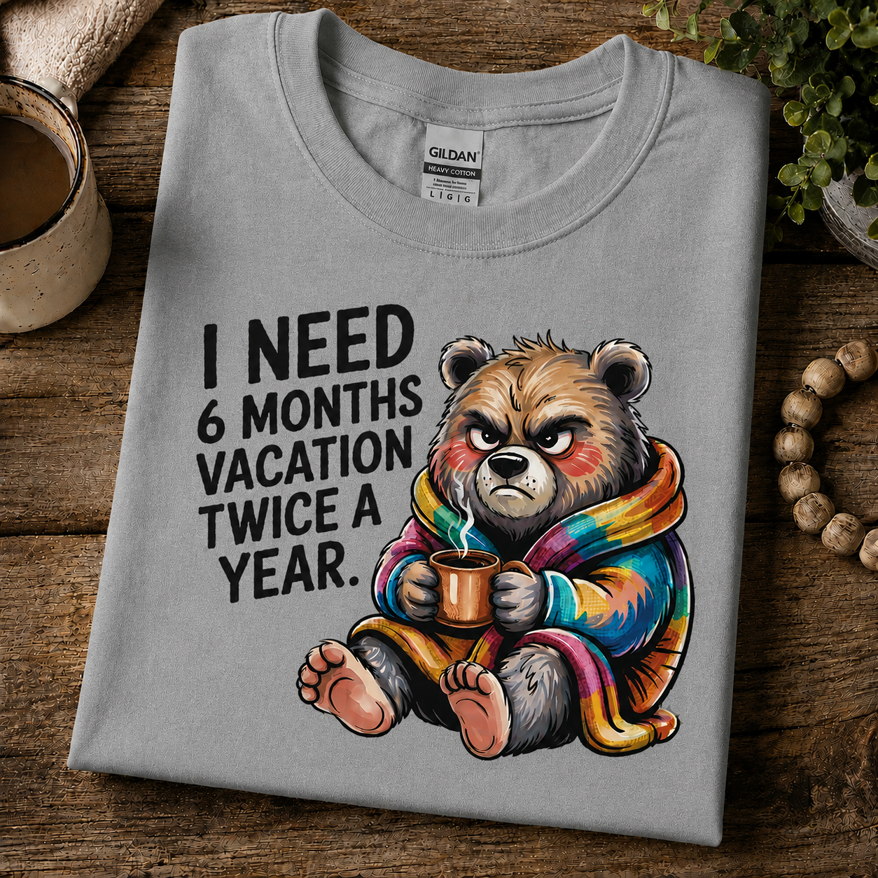 A folded sport gray Gildan t-shirt with a humorous graphic of a grumpy cartoon bear wearing a rainbow blanket and holding a mug, next to the text 'I NEED 6 MONTHS VACATION TWICE A YEAR.' The shirt is styled on a rustic wooden table with a coffee cup, plant, and wooden beads.