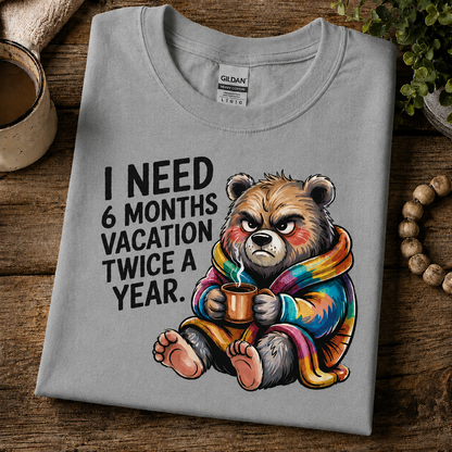 A folded sport gray Gildan t-shirt with a humorous graphic of a grumpy cartoon bear wearing a rainbow blanket and holding a mug, next to the text 'I NEED 6 MONTHS VACATION TWICE A YEAR.' The shirt is styled on a rustic wooden table with a coffee cup, plant, and wooden beads.