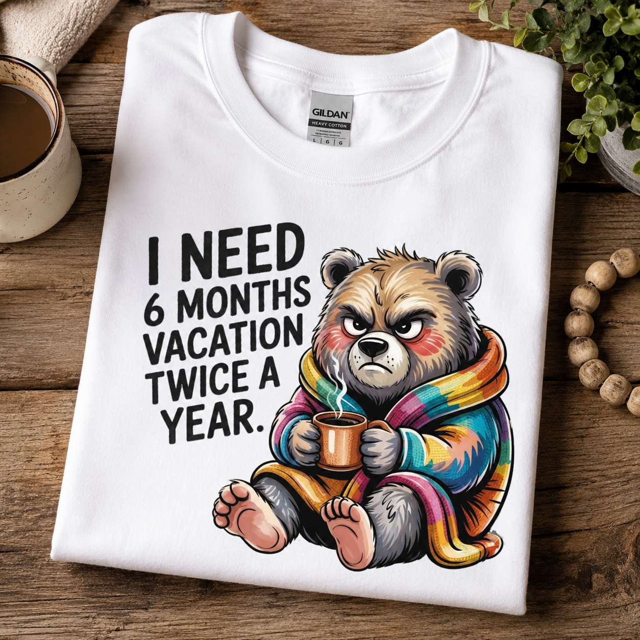 A folded white Gildan t-shirt with a humorous graphic of a grumpy bear wrapped in a rainbow blanket, sitting on a rustic wooden table next to a mug and beads.