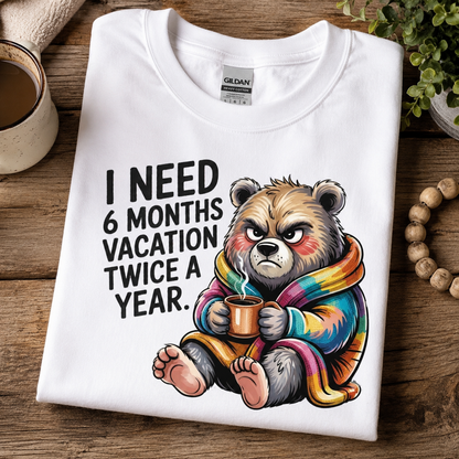 A folded white Gildan t-shirt with a humorous graphic of a grumpy bear wrapped in a rainbow blanket, sitting on a rustic wooden table next to a mug and beads.