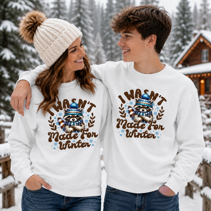 A man and a woman standing together in a snowy outdoor setting, both wearing matching white crewneck sweatshirts with a brown raccoon graphic and the text 'I WASN'T Made FOR WINTER' in brown lettering, along with blue and brown accents.