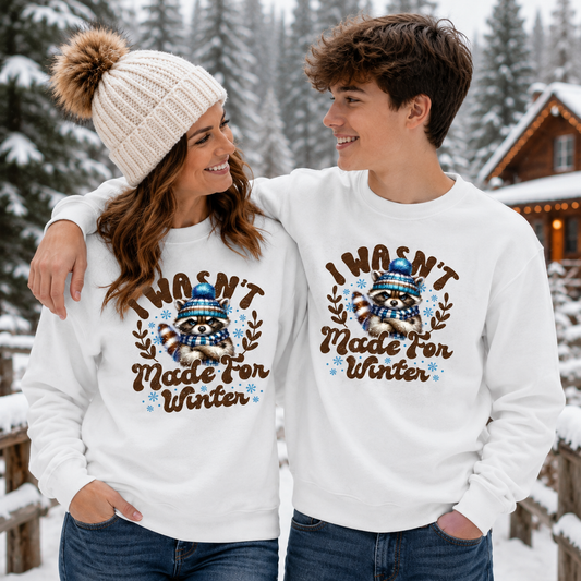 A man and a woman standing together in a snowy outdoor setting, both wearing matching white crewneck sweatshirts with a brown raccoon graphic and the text 'I WASN'T Made FOR WINTER' in brown lettering, along with blue and brown accents.
