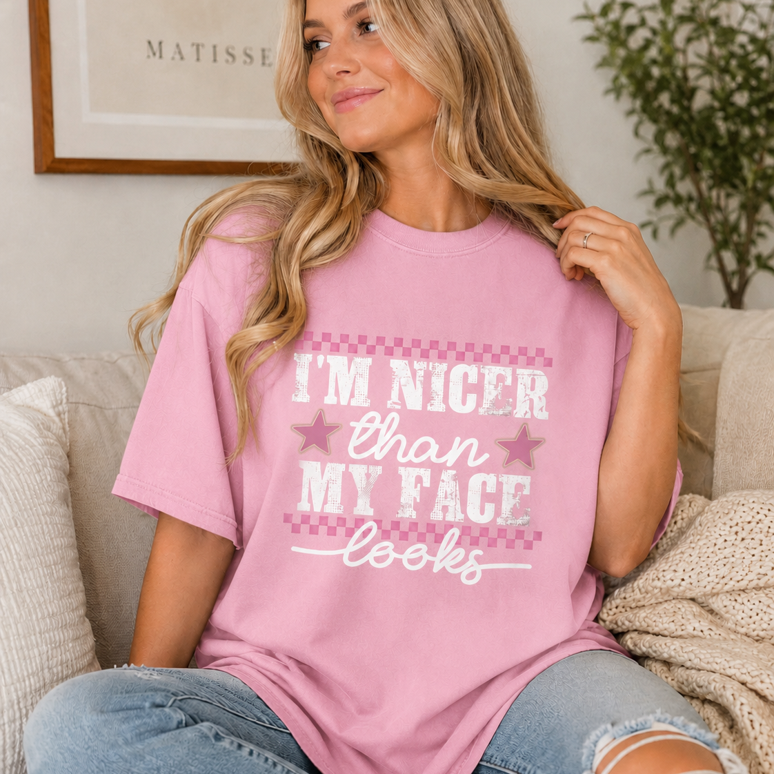 Woman with long blonde hair sitting on a couch wearing a blossom (pink) graphic t-shirt that says “I’m Nicer Than My Face Looks”