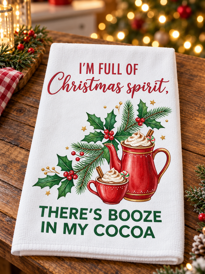 A white kitchen towel with a festive Christmas print, featuring the text 'I'M FULL OF Christmas spirit, THERE'S BOOZE IN MY COCOA' along with illustrations of a red mug, hot cocoa, and holly. The towel is displayed on a rustic wooden table with holiday decorations in the background.