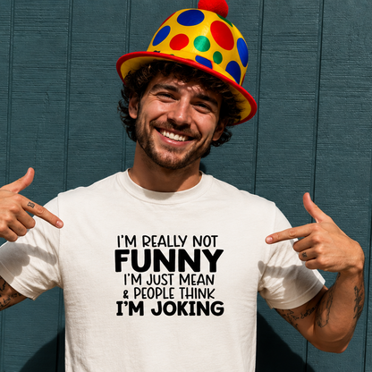 Man wearing a colorful clown hat and a white t-shirt with humorous text, pointing at himself against a blue wall.