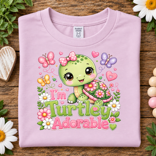 A folded ballerina pink t-shirt with a colorful cartoon turtle graphic and the text 'I'm Turtley Adorable' on a rustic wooden background decorated with flowers and a heart.