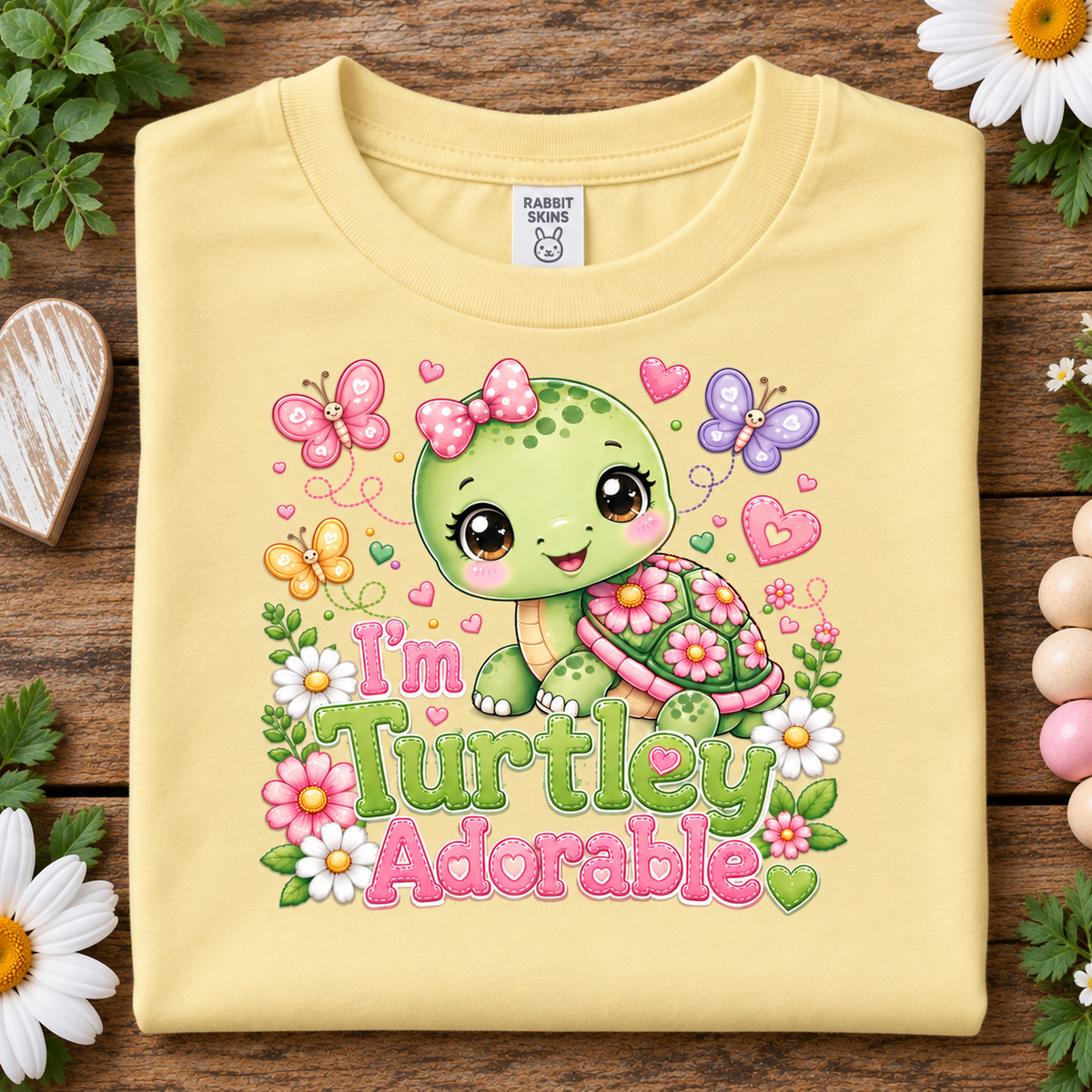 A butter yellow folded T-shirt laid flat on a rustic wooden surface, surrounded by flowers and greenery. The shirt features a colorful cartoon turtle graphic with butterflies, hearts, flowers, and the text 'I'm Turtley Adorable.