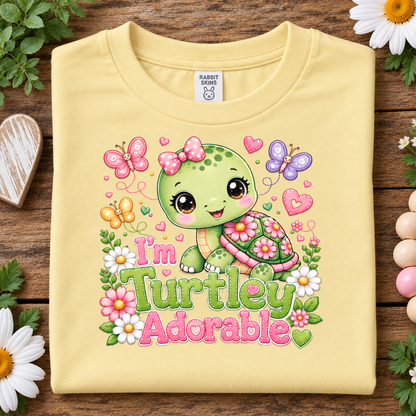 A butter yellow folded T-shirt laid flat on a rustic wooden surface, surrounded by flowers and greenery. The shirt features a colorful cartoon turtle graphic with butterflies, hearts, flowers, and the text 'I'm Turtley Adorable.