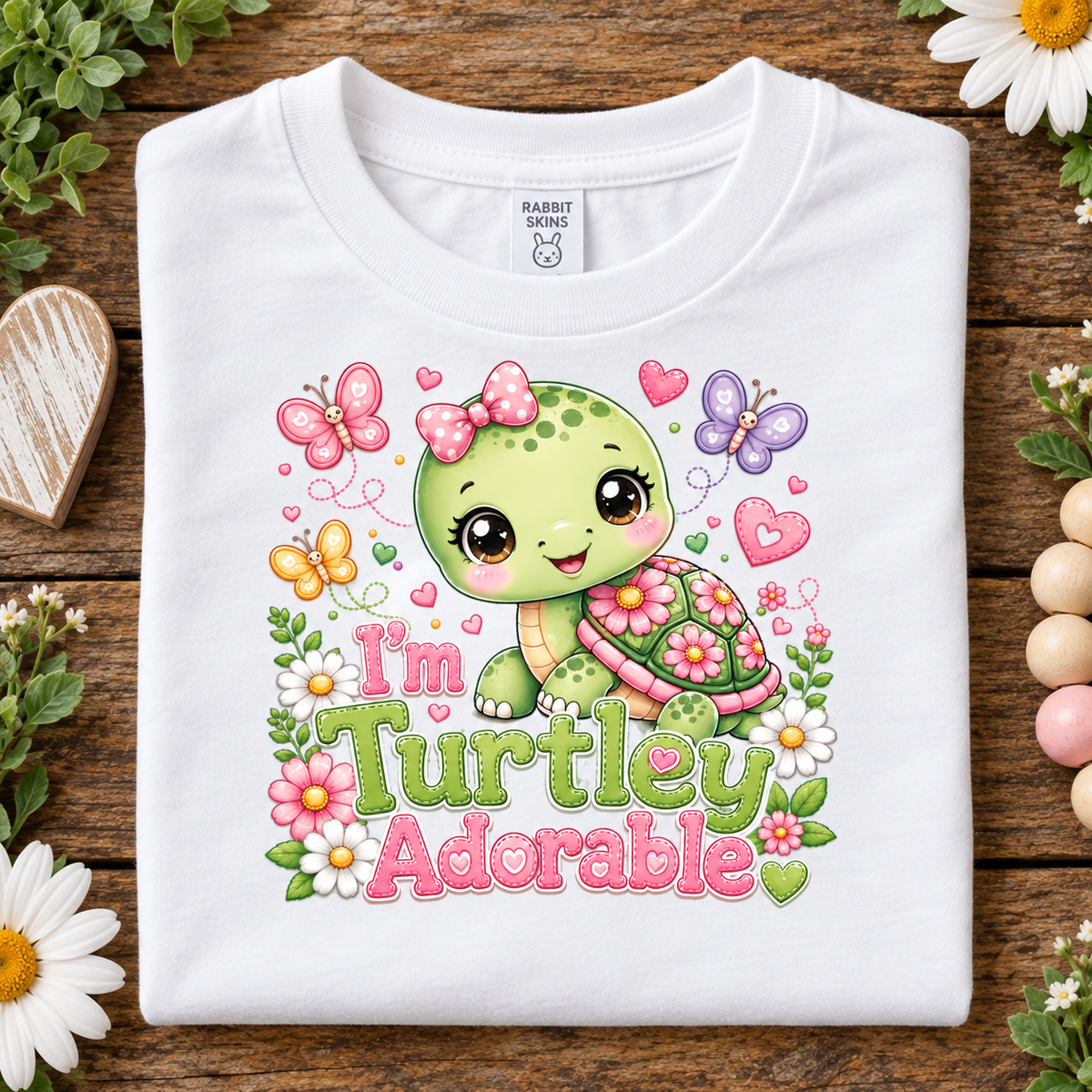 A folded white short-sleeve T-shirt with a colorful cartoon turtle graphic and the text 'I'm Turtley Adorable' on a rustic wooden surface surrounded by flowers and decorative props.