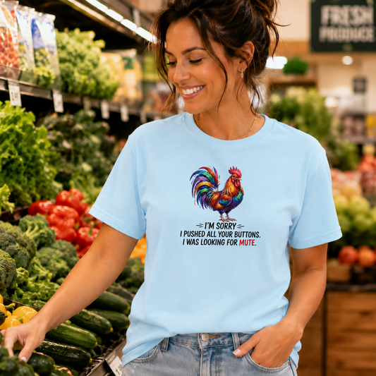 A woman in a grocery store wearing a light blue graphic t-shirt with a colorful rooster and the text 'I'M SORRY I PUSHED ALL YOUR BUTTONS. I WAS LOOKING FOR MUTE.
