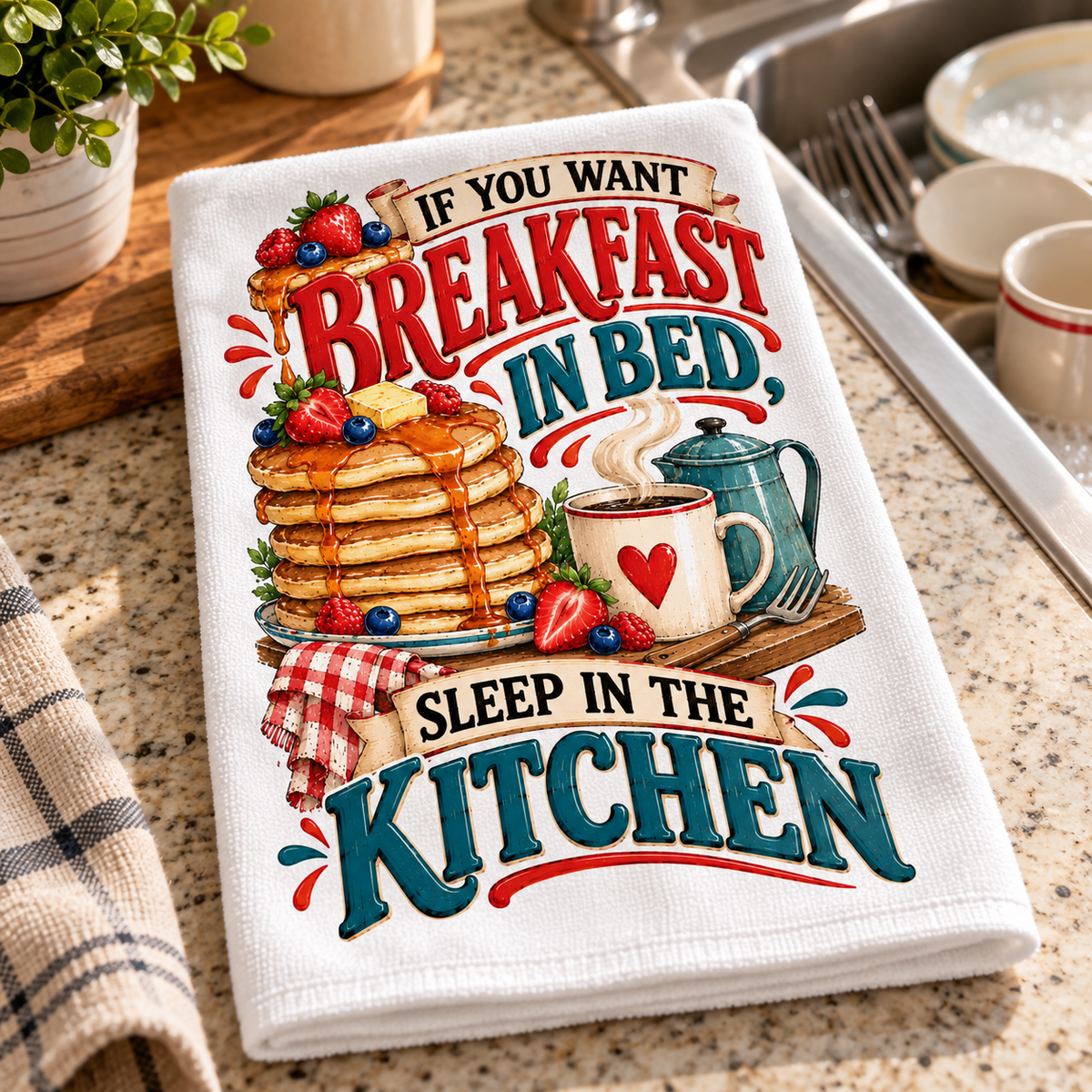 A folded white kitchen towel with a colorful breakfast-themed graphic reading 'IF YOU WANT BREAKFAST IN BED. SLEEP IN THE KITCHEN.