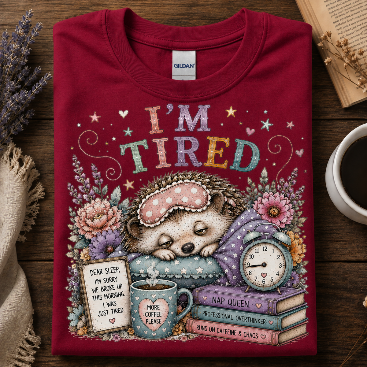 A folded cardinal red Gildan t-shirt with a colorful graphic of a sleeping hedgehog surrounded by flowers, a clock, books, and coffee mugs, with the text 'I'M TIRED'.