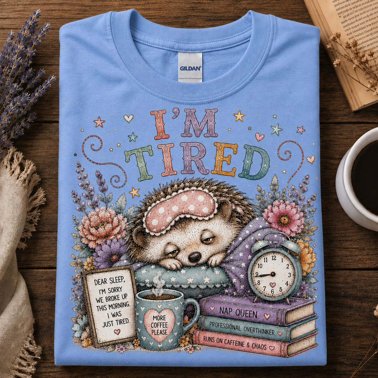 A carolina blue Gildan t-shirt with a detailed graphic of a sleeping hedgehog surrounded by cozy elements like a mug, alarm clock, and books, with the text 'I'M TIRED'. The shirt is displayed flat on a wooden surface with decorative props.