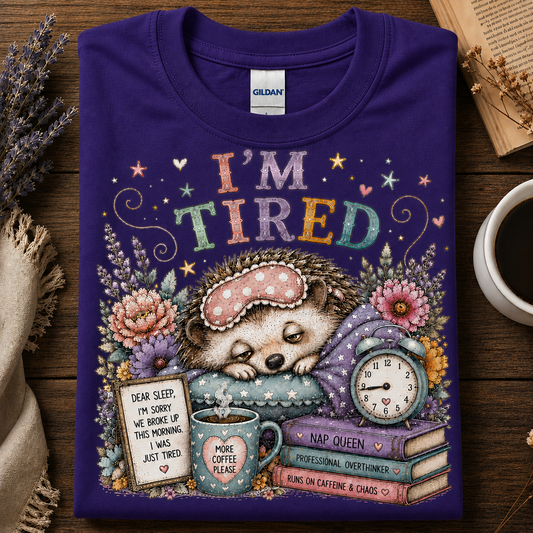 A folded purple Gildan t-shirt with a colorful graphic of a sleeping hedgehog under the text 'I'M TIRED', surrounded by flowers, a mug, a clock, and books, displayed on a wooden surface with decorative props.