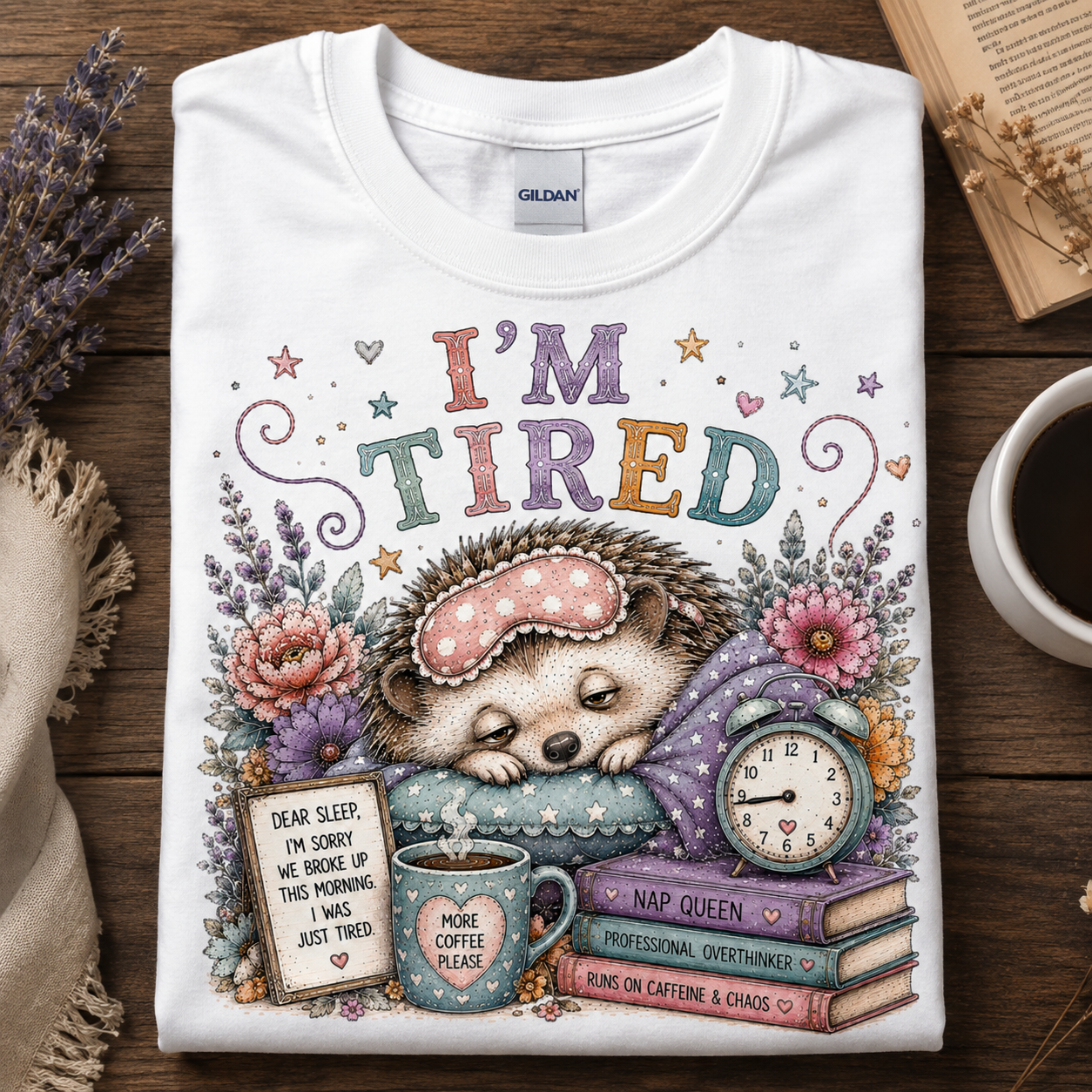 A folded white Gildan t-shirt with a colorful 'I'M TIRED' hedgehog graphic, displayed flat on a wooden table with a coffee cup, dried lavender, and an open book.
