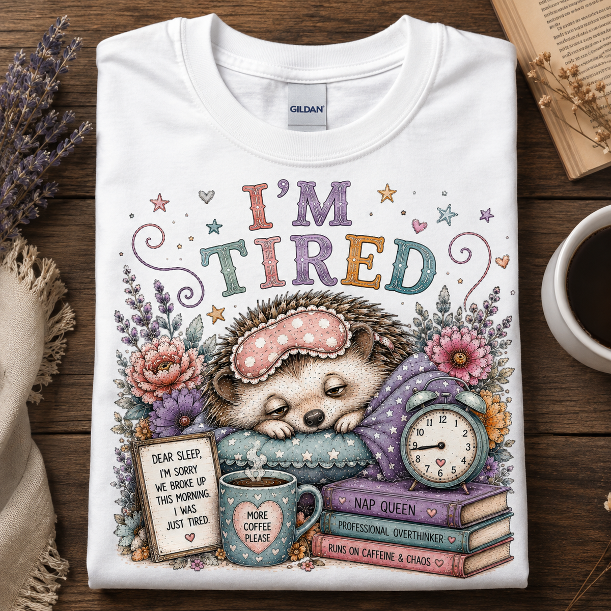 A folded white Gildan t-shirt with a colorful 'I'M TIRED' hedgehog graphic, displayed flat on a wooden table with a coffee cup, dried lavender, and an open book.