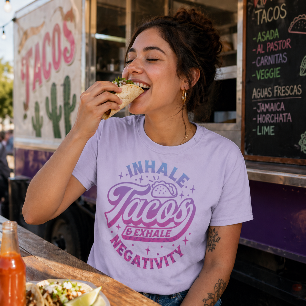 Woman eating a taco in front of a taco truck wearing a lilac colored shirt with a taco-themed message.