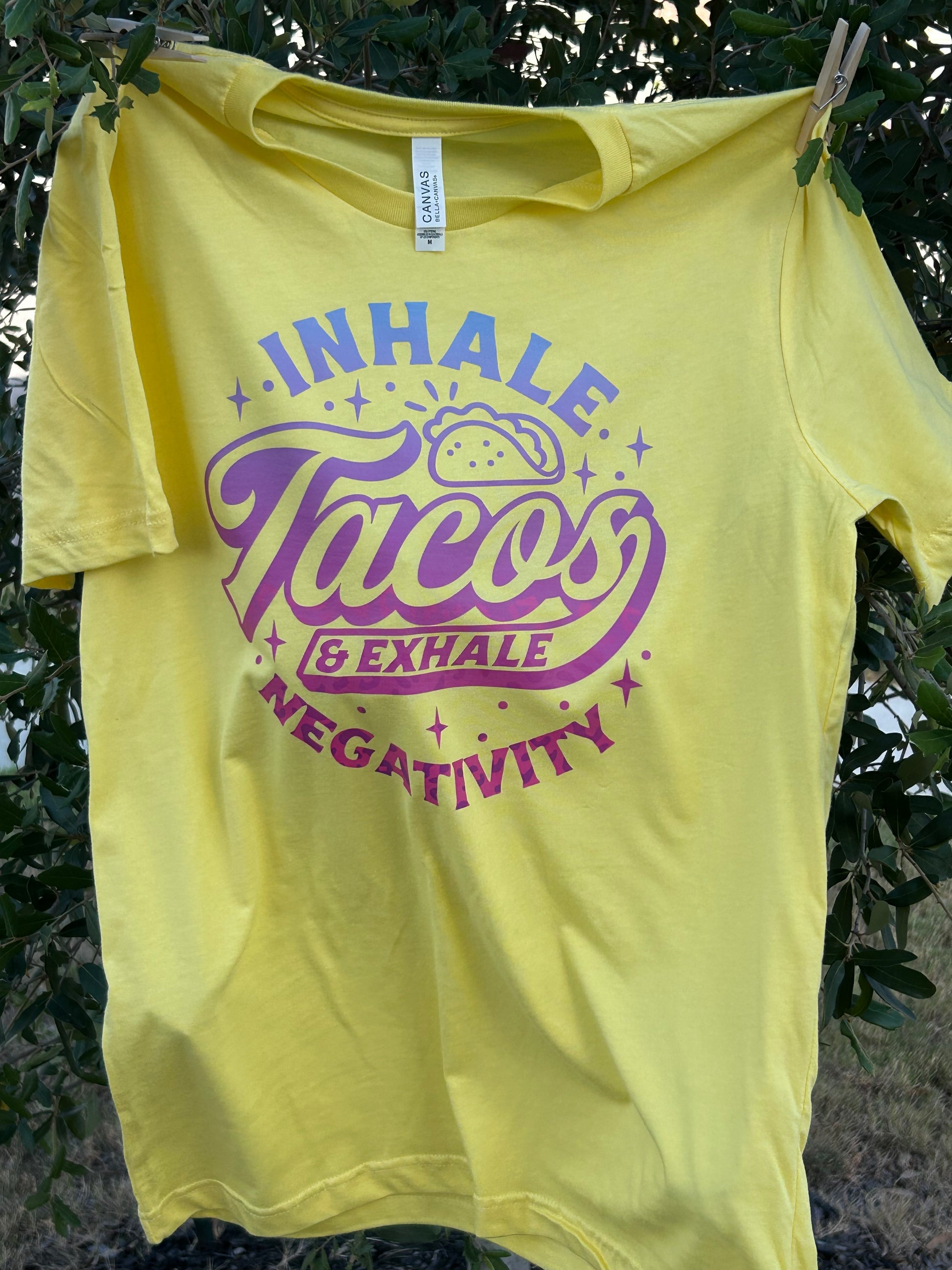 Yellow t-shirt with purple graphic and text 'INHALE TACOS BREATHE NEGATIVELY' hanging on a tree branch.