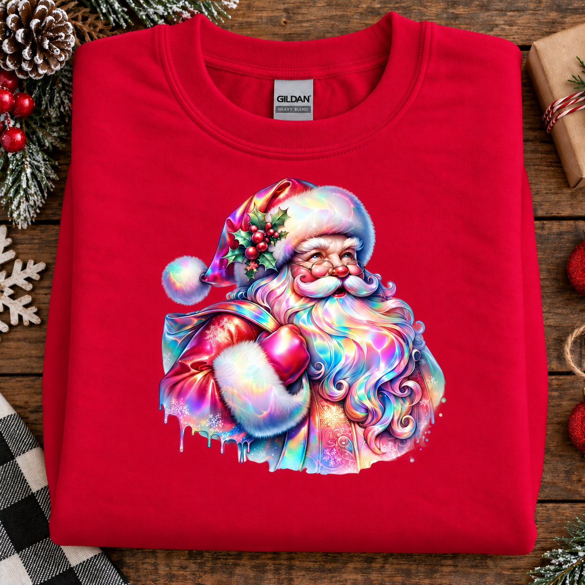 A folded red Gildan sweatshirt with a colorful, rainbow-iridescent graphic of Santa Claus on the front, styled on a rustic wooden surface with holiday decorations.