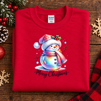 A folded red Gildan Youth crewneck sweatshirt with a colorful watercolor snowman graphic and 'Merry Christmas' text, displayed on a rustic wooden background with holiday decorations.