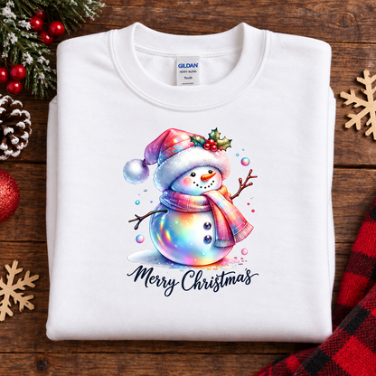 A folded white Gildan sweatshirt with a colorful Christmas snowman graphic and 'Merry Christmas' text, displayed on a rustic wooden background with holiday decorations.