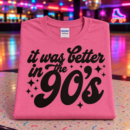 A folded azalea pink Gildan Softstyle t-shirt with a black graphic reading 'it was better in the 90's' sits on a colorful, speckled tabletop with a blurred roller skate arcade background.