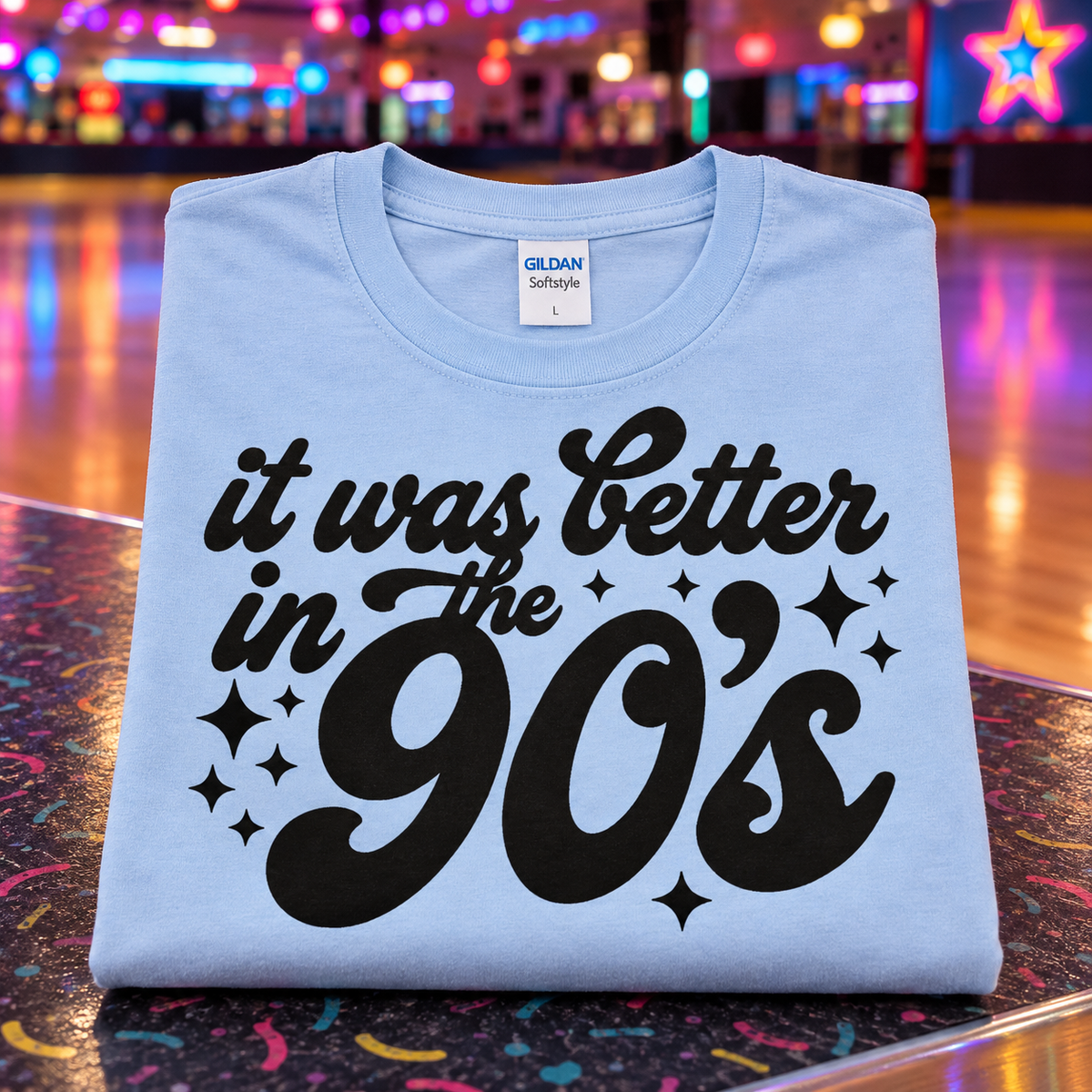 A folded light blue Gildan Softstyle t-shirt with a black graphic reading 'it was better in the 90's' and star accents, displayed on a colorful, confetti-covered table with a blurred roller rink background.