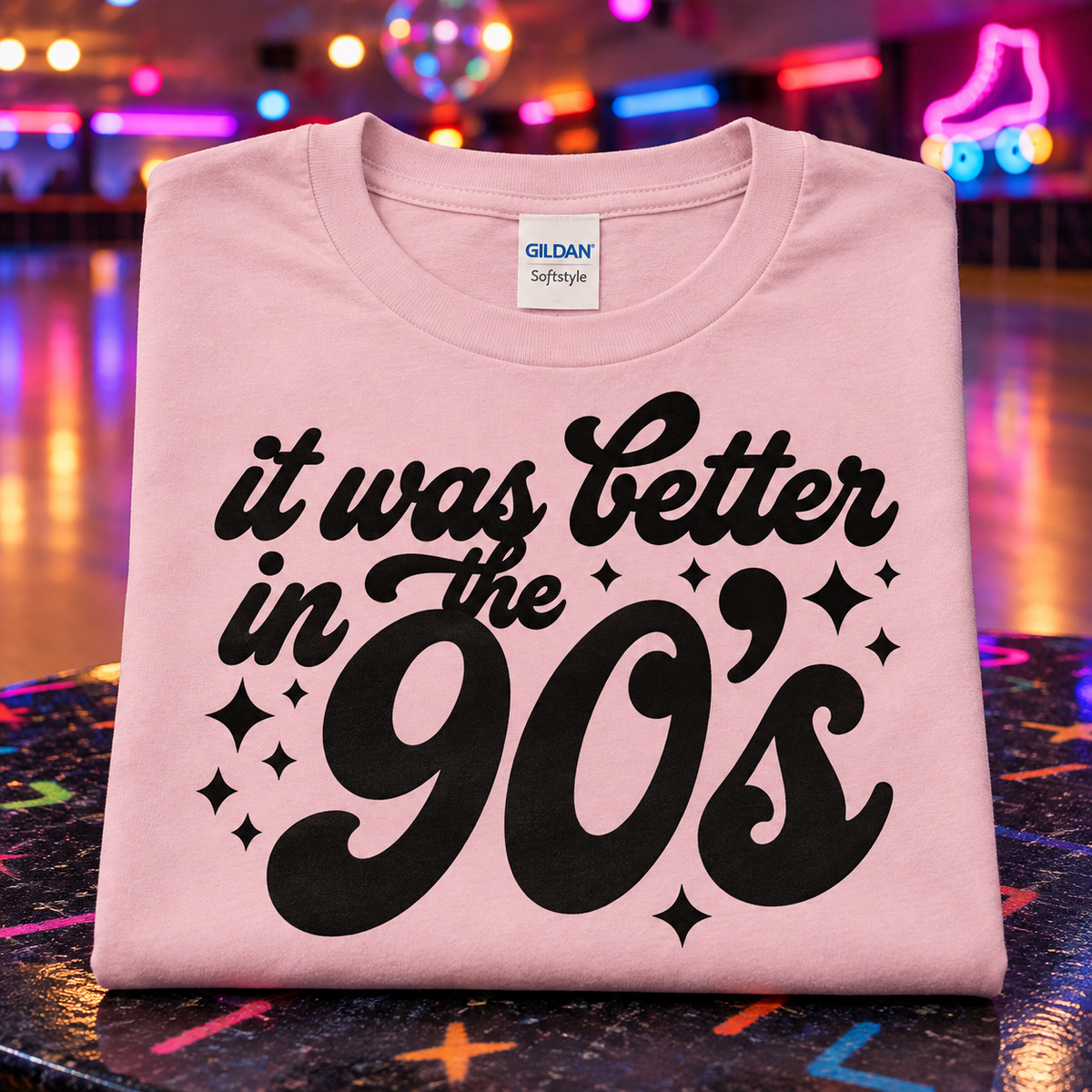A folded light pink Gildan Softstyle t-shirt with a black graphic reading 'it was Better in the 90's' and star accents, displayed on a colorful roller-skating rink floor with neon lights in the background.