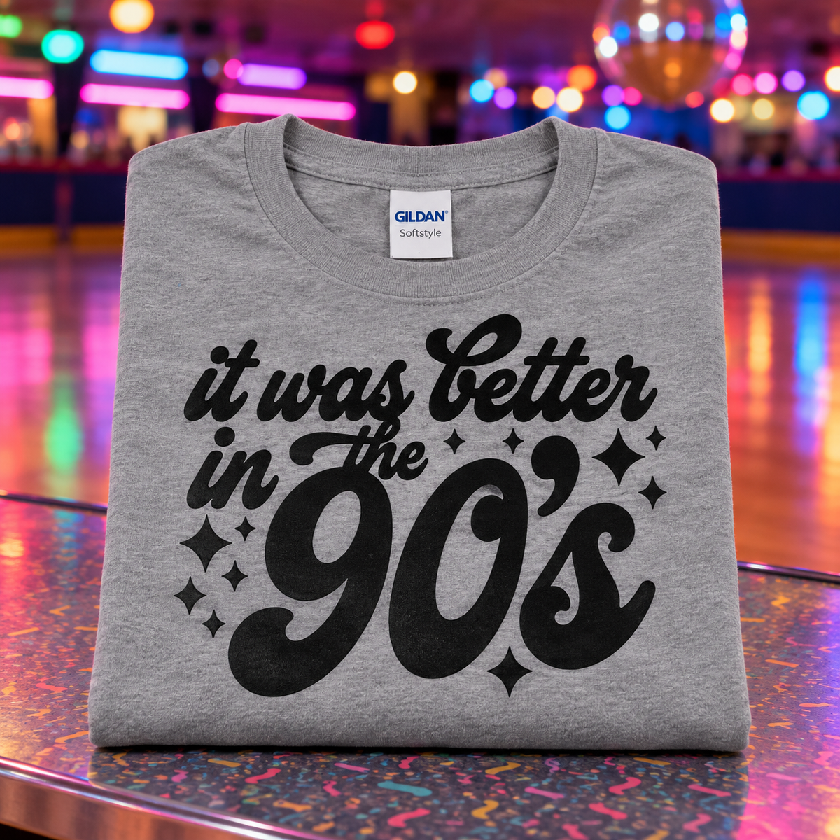 A folded heather gray Gildan Softstyle t-shirt with a black graphic reading 'it was better in the 90's' and sparkles, displayed on a colorful, reflective surface with a blurred neon-lit background.