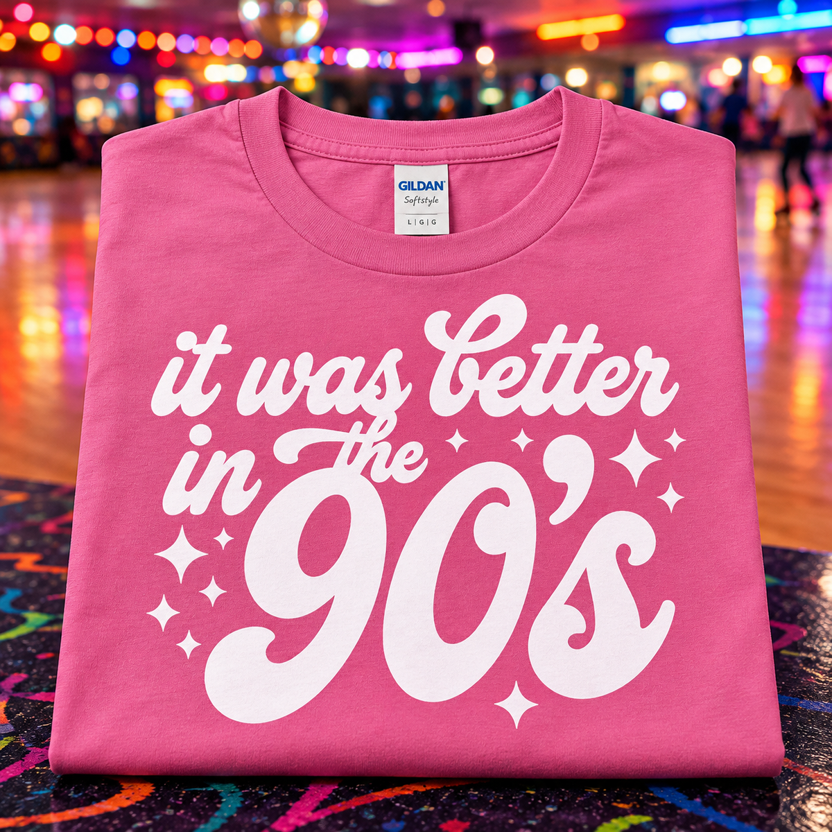 A folded azalea pink Gildan t-shirt with a white graphic reading 'it was better in the 90's' and sparkles, displayed against a blurred, colorful indoor amusement park background.