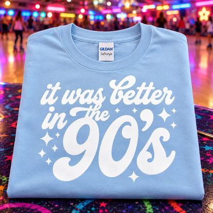 A folded light blue Gildan Softstyle t-shirt with a white graphic reading 'it was Better in the 90's' and sparkles, displayed on a colorful, starry surface with a blurred amusement park background.