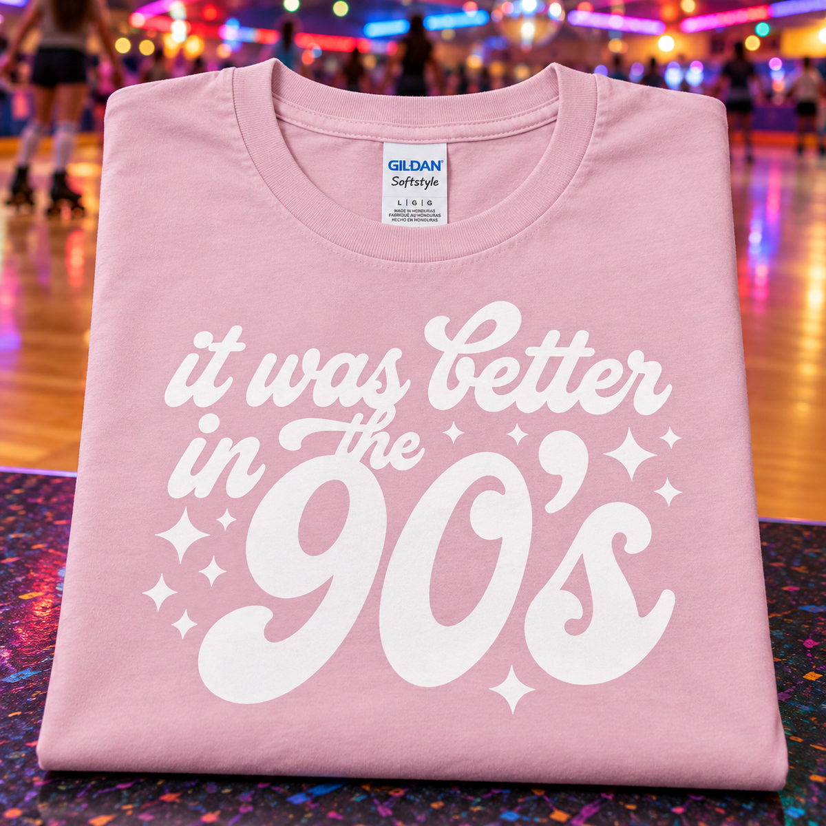 A folded light pink Gildan Softstyle t-shirt with a white retro graphic reading 'it was better in the 90's' and star accents, displayed on a colorful, glittery surface with a blurred roller rink background.