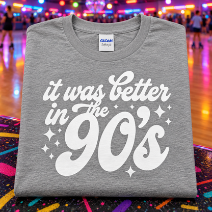 A folded heather gray Gildan Comfortstyle t-shirt with a white graphic reading 'it was better in the 90's' and sparkles, displayed on a colorful, patterned surface with a blurred roller rink background.