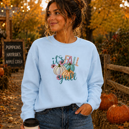 A woman wearing a light blue crewneck sweatshirt with a colorful 'it's fall y'all' graphic featuring pumpkins, standing outdoors in a fall-themed setting with pumpkins and autumn foliage.