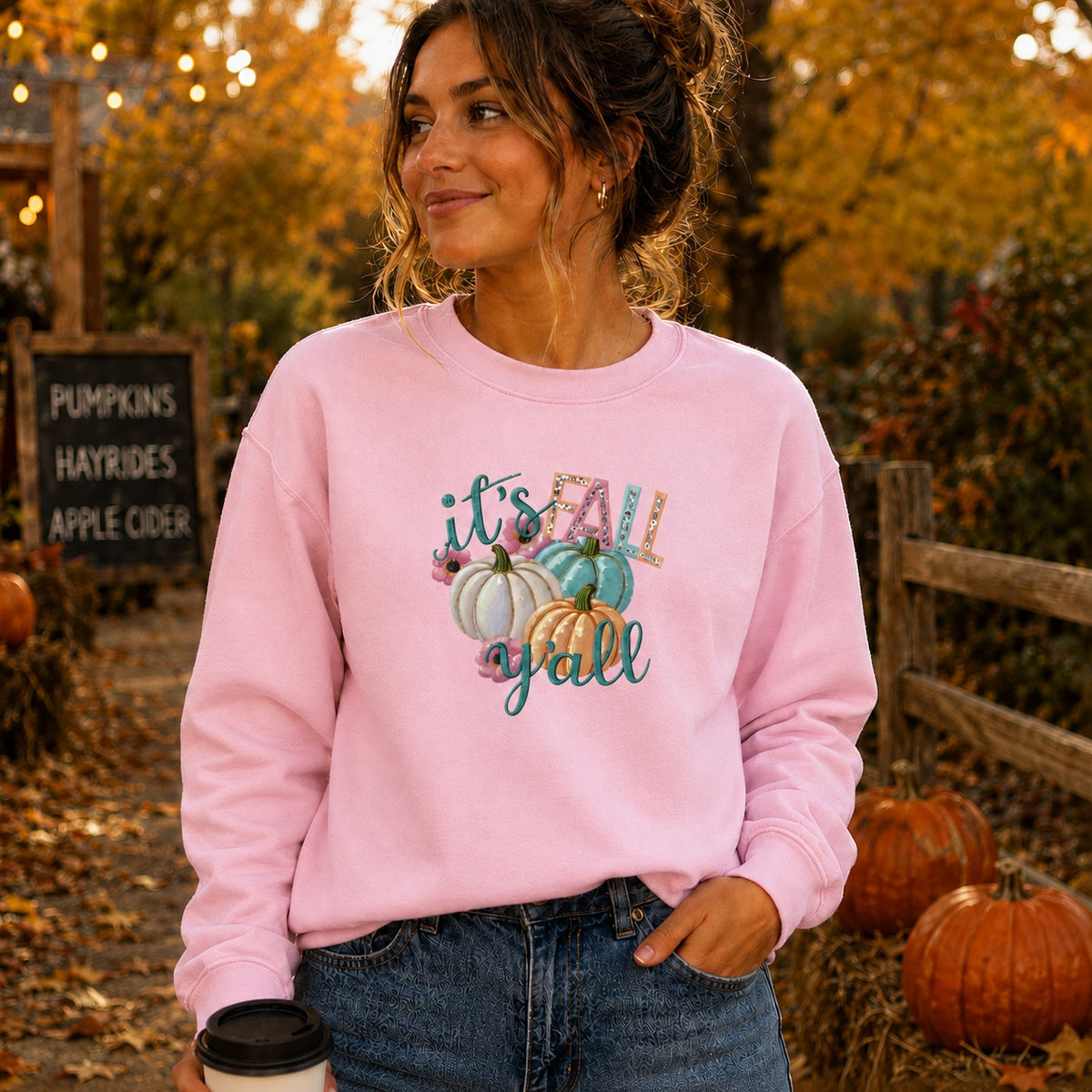 A woman wearing a light pink crewneck sweatshirt with a colorful 'it's all y'all' autumn graphic featuring pumpkins, standing outdoors in a fall garden setting with pumpkins and a sign in the background.