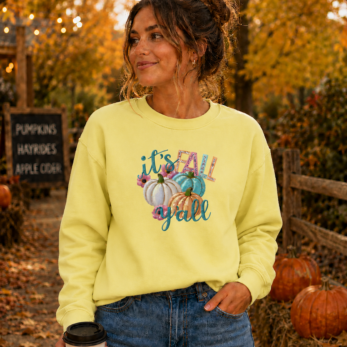 Woman wearing a yellow sweatshirt with fall-themed text and pumpkins, standing in an autumn setting with pumpkins and foliage.