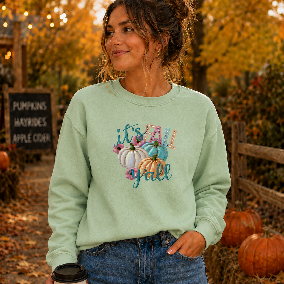 Woman wearing a light green sweatshirt with pumpkin design and text, standing in an autumn setting with pumpkins and foliage.