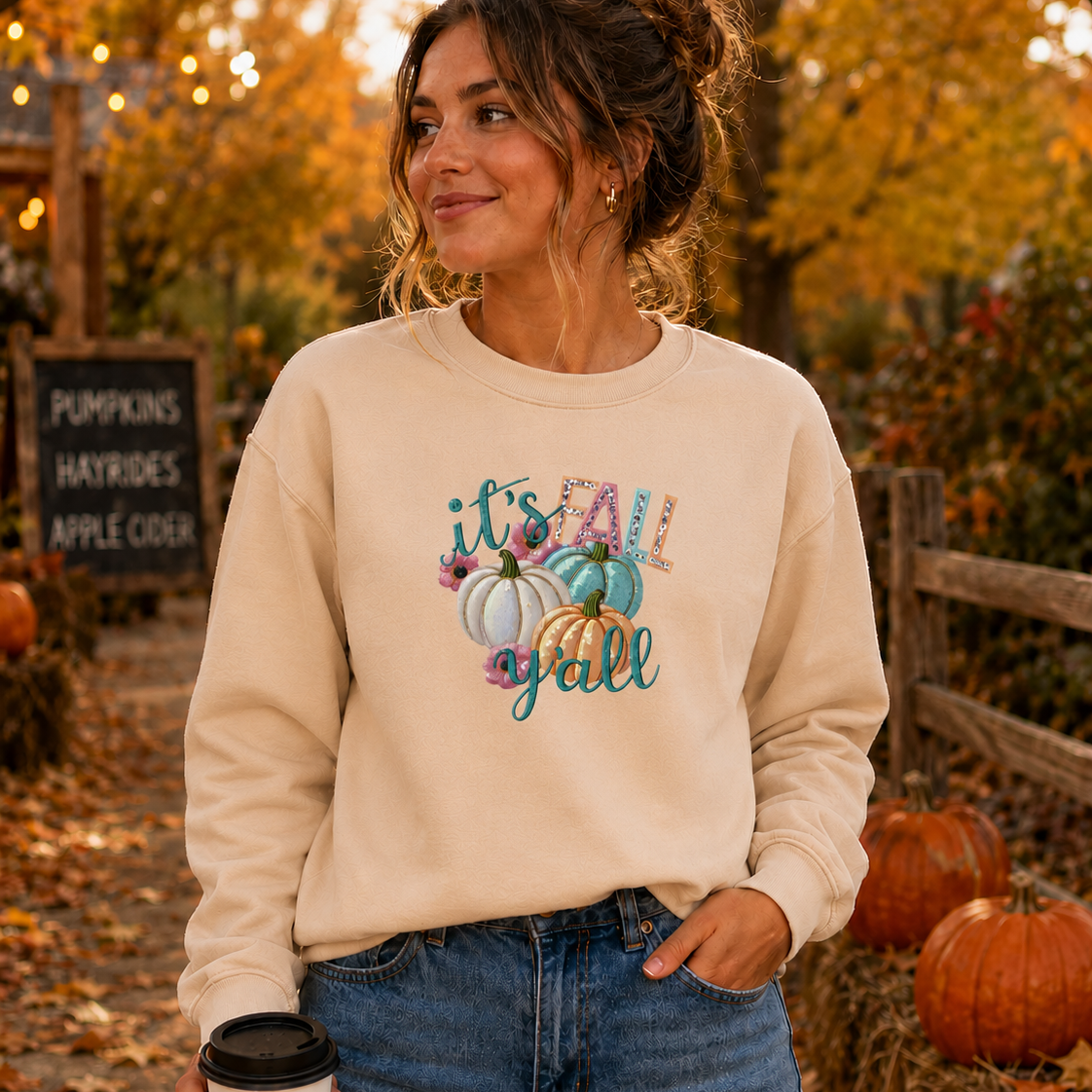 Woman wearing a beige sweatshirt with a fall-themed design, standing in an autumn setting with pumpkins and foliage.