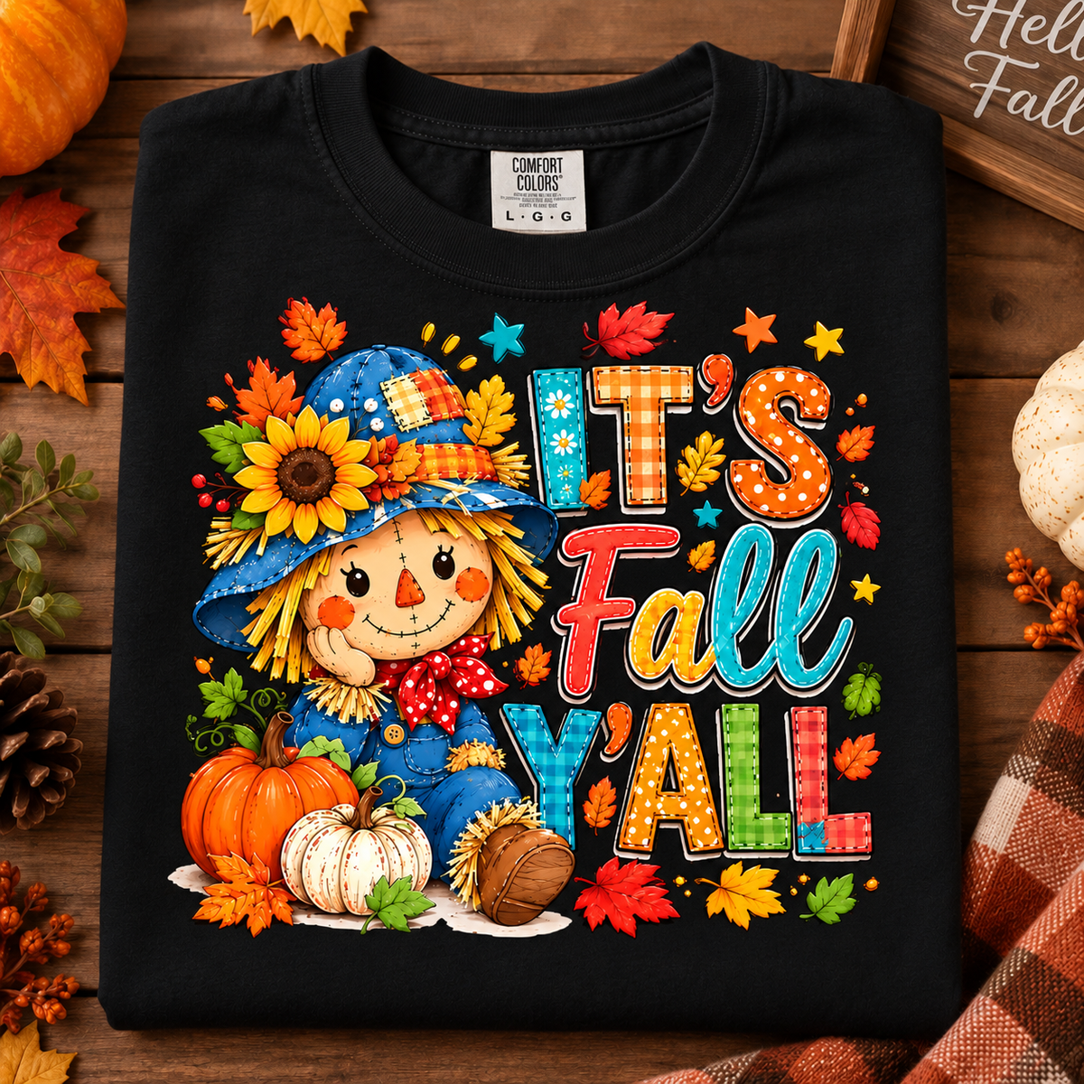 A black Comfort Colors t-shirt with a colorful 'It's Fall Y'all' autumn graphic featuring a scarecrow, pumpkins, and fall leaves, styled on a wooden background with seasonal props.