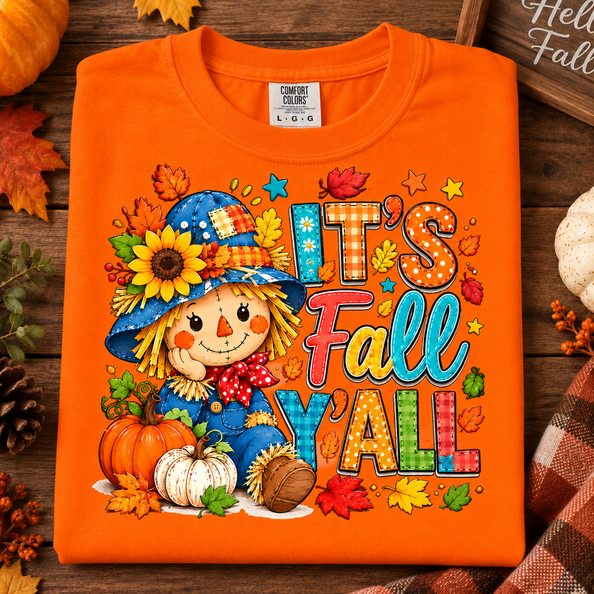 An burnt orange short-sleeve t-shirt with a colorful 'It's Fall Y'all' autumn scarecrow graphic, displayed on a rustic wooden background with fall-themed props.