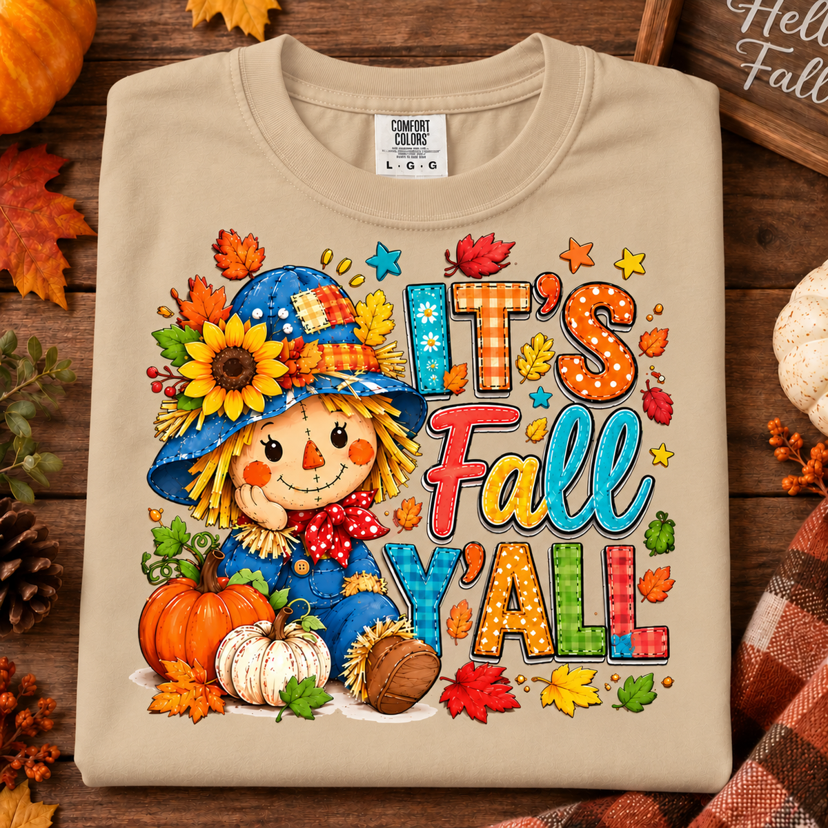 A sandstone crewneck t-shirt with a colorful 'It's Fall Y'all' graphic featuring a cartoon scarecrow, pumpkins, and autumn leaves, styled in a flat lay on a wooden surface with fall decorations.