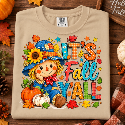 A sandstone crewneck t-shirt with a colorful 'It's Fall Y'all' graphic featuring a cartoon scarecrow, pumpkins, and autumn leaves, styled in a flat lay on a wooden surface with fall decorations.